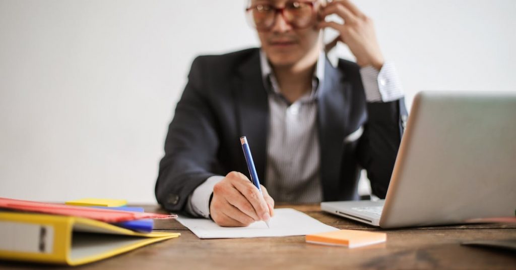 Businessman talking on phone while writing notes and using a laptop at desk.