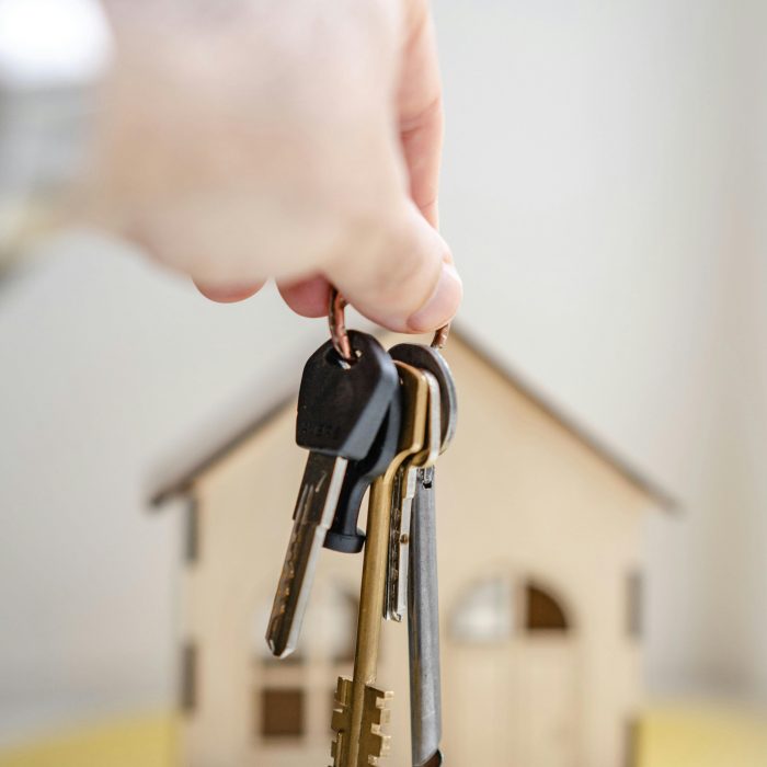 Startseite Close-up of a hand holding keys with a miniature wooden house in the background, symbolizing real estate investment.